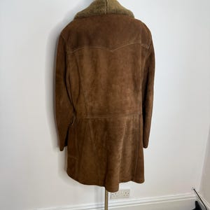 May include: A brown suede coat with a faux fur collar is displayed on a mannequin. The coat has a classic design with a tailored fit, featuring a notched collar and a button-up front. The coat is a warm brown colour.