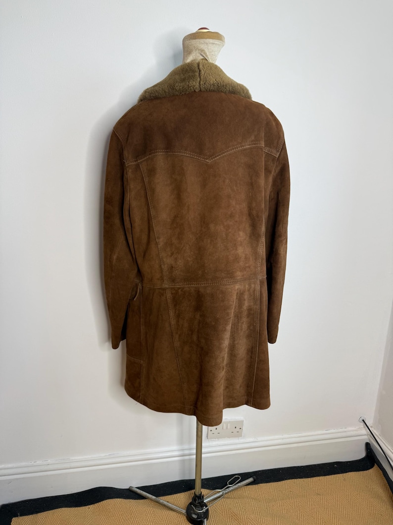 May include: A brown suede coat with a faux fur collar is displayed on a mannequin. The coat has a classic design with a tailored fit, featuring a notched collar and a button-up front. The coat is a warm brown colour.