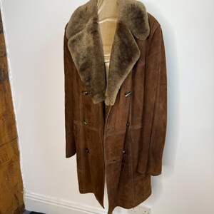 May include: A brown suede coat with a shearling collar and lining. The coat features a double-breasted design with large buttons and a knee-length cut. The coat is displayed on a mannequin.