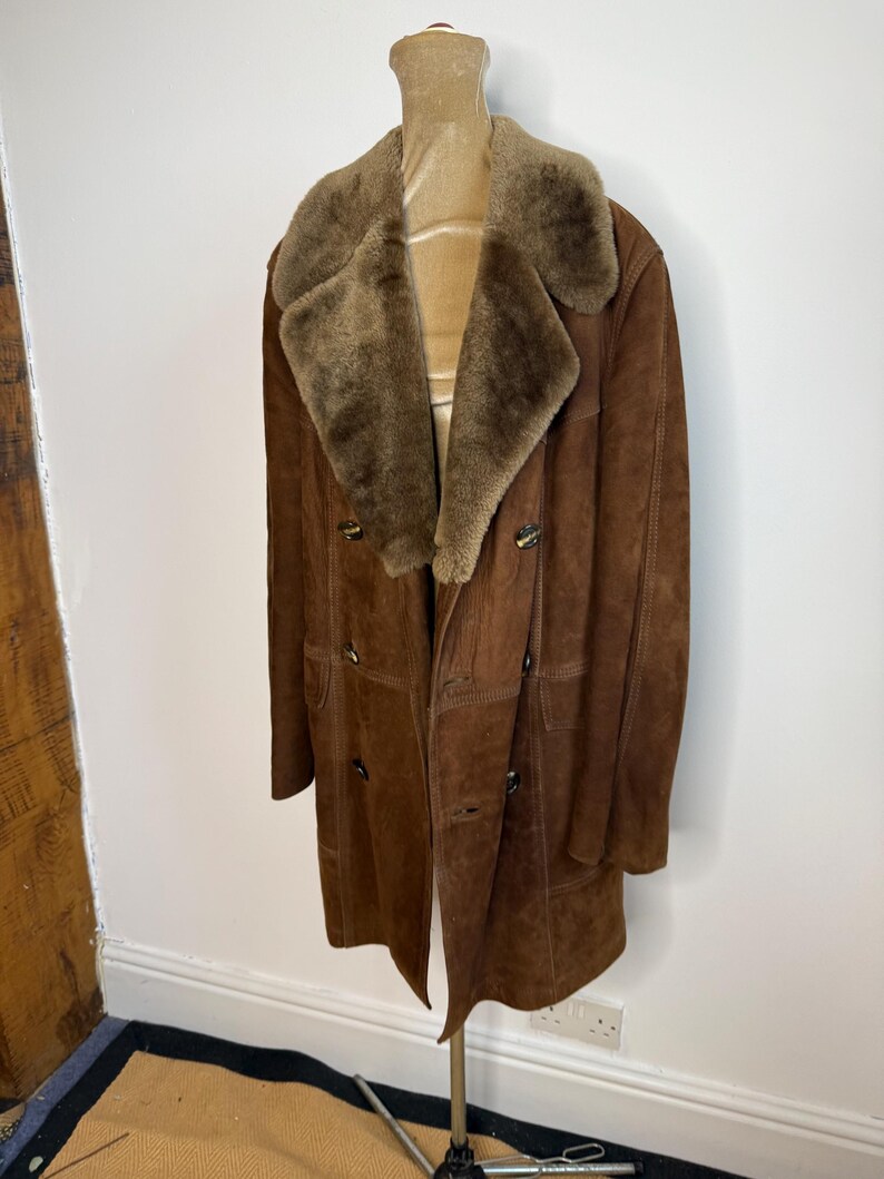 May include: A brown suede coat with a shearling collar and lining. The coat features a double-breasted design with large buttons and a knee-length cut. The coat is displayed on a mannequin.