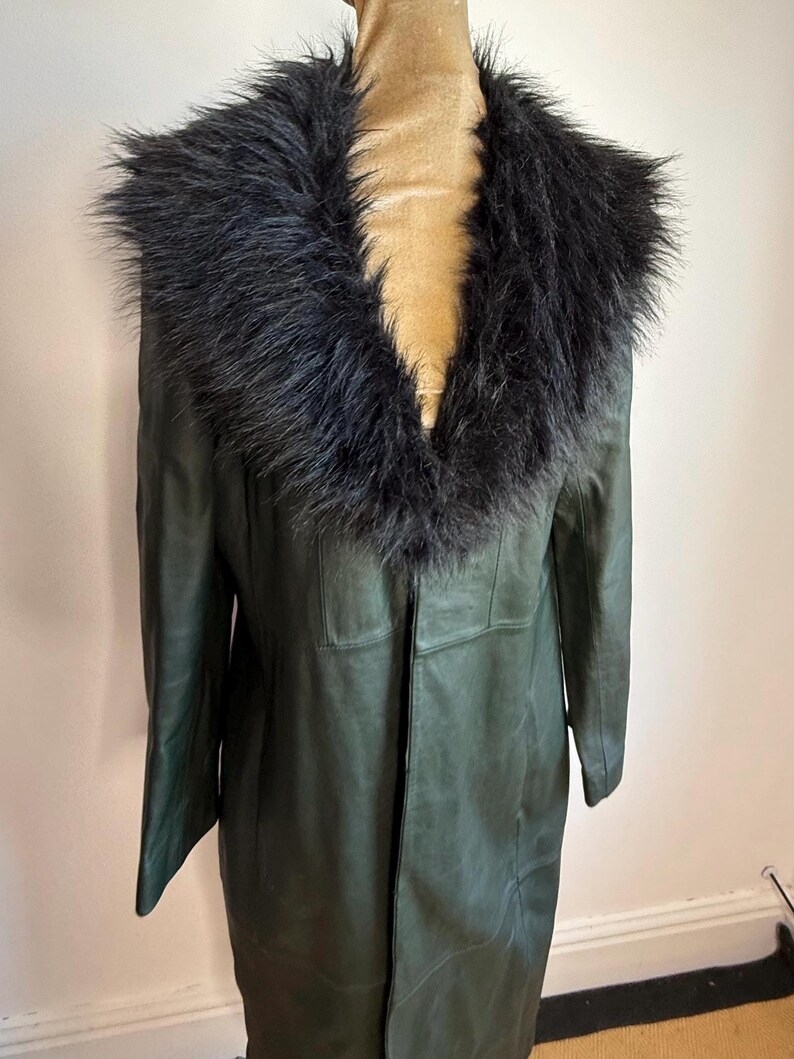 May include: A dark green leather coat with a black faux fur collar. The coat is long, reaching the mid-calf, and has a classic, tailored design. The collar adds a touch of luxury to the outerwear.