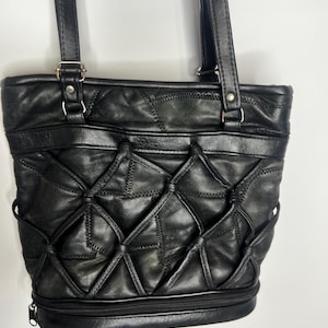 Puede incluir: Un bolso tote de cuero negro con un revestimiento de tiras de cuero en forma de celosía en el panel frontal. El bolso incluye dos correas para el hombro sujetas con herrajes de metal plateado y un compartimento horizontal con cremallera en la base.