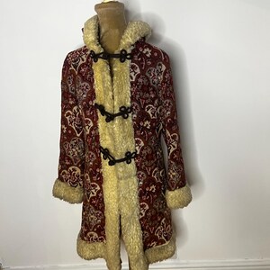 Puede incluir: Un abrigo rojo vintage con un estampado floral, ribete de piel sintética color crema y cierres de palanca. El abrigo tiene capucha y mangas largas. El abrigo se exhibe en un maniquí.