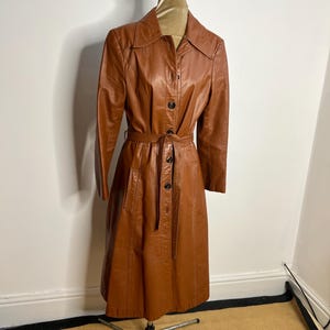 May include: A vintage brown leather trench coat displayed on a mannequin. The coat features a collared neckline, button-down front, and a matching belt. The coat has long sleeves and pockets. The coat is knee-length.