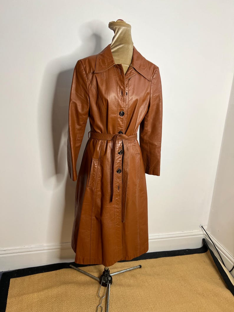 May include: A vintage brown leather trench coat displayed on a mannequin. The coat features a collared neckline, button-down front, and a matching belt. The coat has long sleeves and pockets. The coat is knee-length.