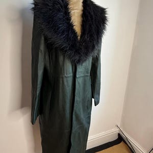 May include: A dark green leather coat with a black faux fur collar. The long coat is displayed on a mannequin. The coat has a classic design with a tailored fit.