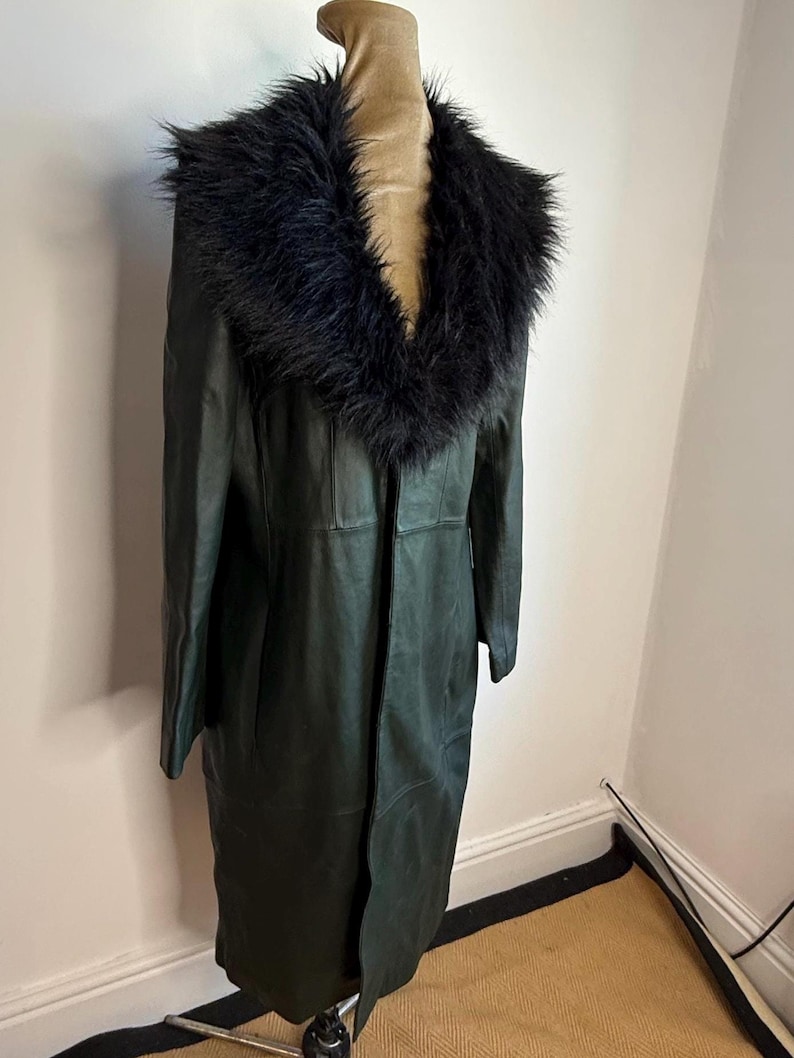 May include: A dark green leather coat with a black faux fur collar. The long coat is displayed on a mannequin. The coat has a classic design with a tailored fit.