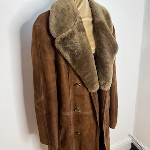 May include: A brown suede coat with a shearling collar and lapels. The double-breasted coat has three buttons and a tailored fit. The coat is displayed on a mannequin. This vintage coat is a classic outerwear piece.