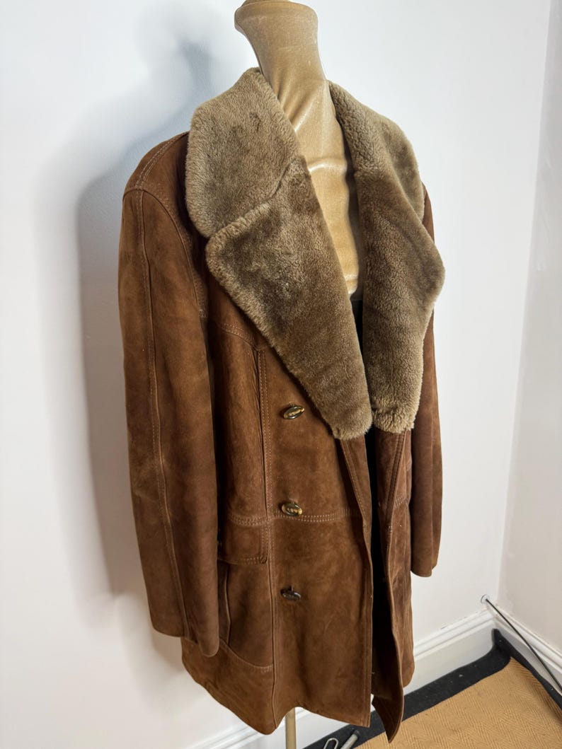 May include: A brown suede coat with a shearling collar and lapels. The double-breasted coat has three buttons and a tailored fit. The coat is displayed on a mannequin. This vintage coat is a classic outerwear piece.