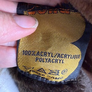 May include: A close-up of a gold tag with black trim, featuring the text "100% ACRYL/ACRYLIQUE POLYACRYL" in white. The tag is attached to a brown, furry material. The tag also has care symbols.