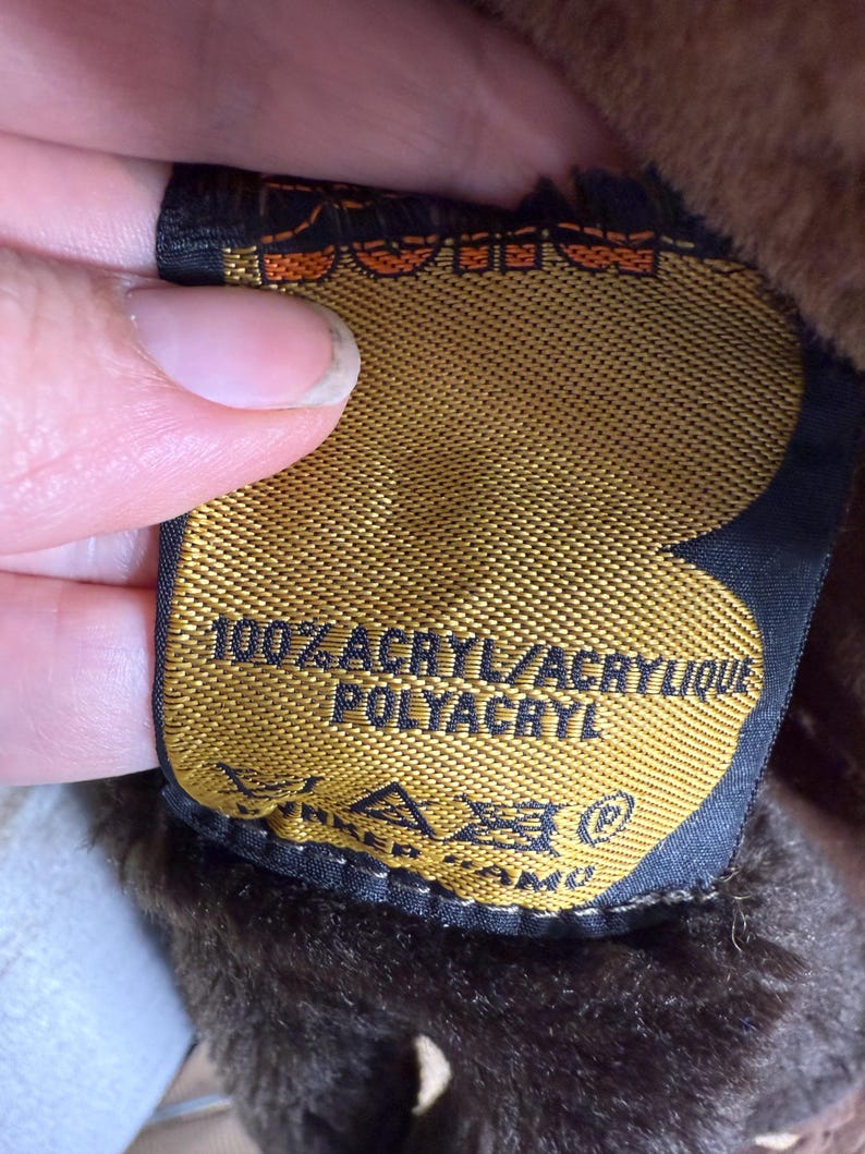 May include: A close-up of a gold tag with black trim, featuring the text "100% ACRYL/ACRYLIQUE POLYACRYL" in white. The tag is attached to a brown, furry material. The tag also has care symbols.