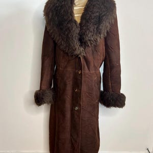 May include: A long, brown suede coat with a fur collar and cuffs. The coat has a button-up front and a classic, vintage style. The fur trim is a darker shade of brown, adding contrast to the coat's overall design.
