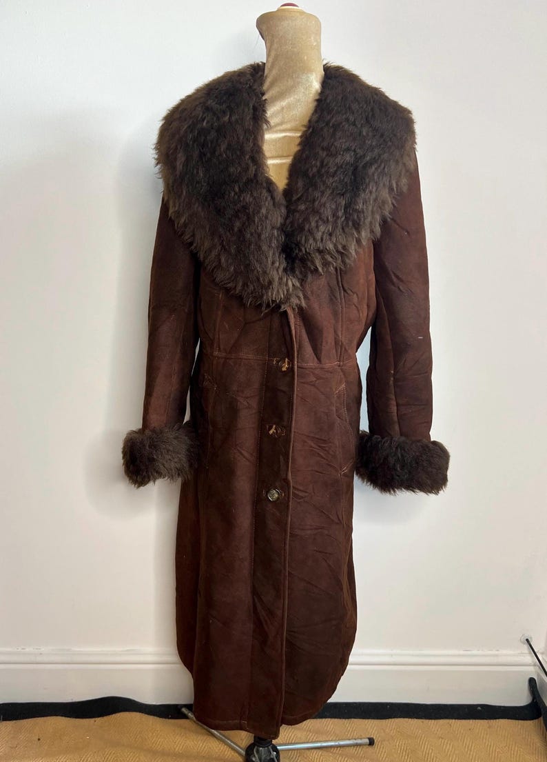May include: A long, brown suede coat with a fur collar and cuffs. The coat has a button-up front and a classic, vintage style. The fur trim is a darker shade of brown, adding contrast to the coat's overall design.