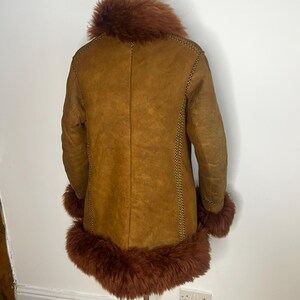 May include: A brown shearling coat with a reddish-brown fur collar and cuffs. The coat has a classic design with visible stitching along the seams. The coat is displayed on a mannequin.