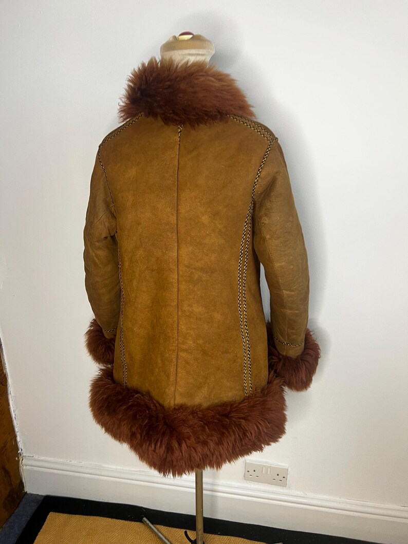 May include: A brown shearling coat with a reddish-brown fur collar and cuffs. The coat has a classic design with visible stitching along the seams. The coat is displayed on a mannequin.