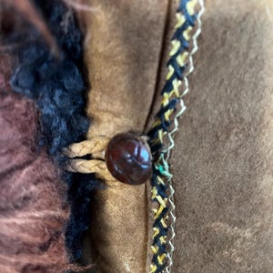 May include: Close-up of a brown suede jacket with a dark brown button and decorative trim. The jacket features a fur collar and a braided leather closure. The trim is black, yellow, and white.