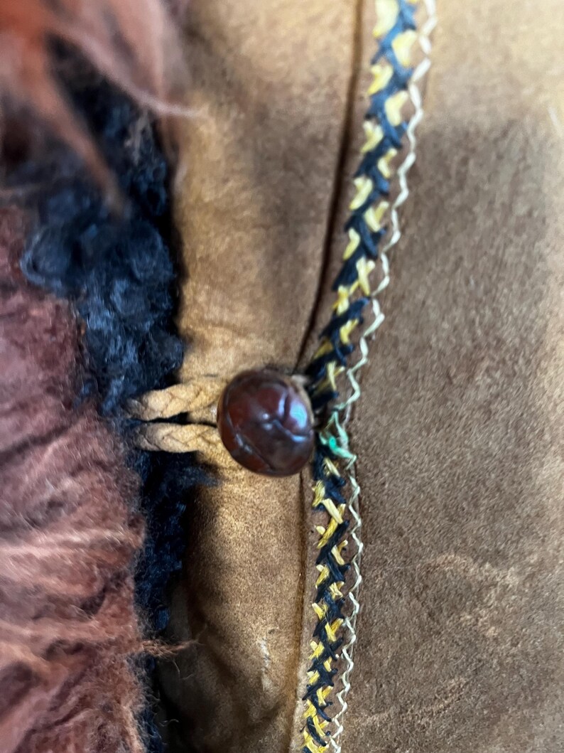 May include: Close-up of a brown suede jacket with a dark brown button and decorative trim. The jacket features a fur collar and a braided leather closure. The trim is black, yellow, and white.