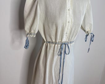 May include: A vintage white dress with a pleated bodice, short sleeves, and a high neckline. The dress features delicate lace trim, small buttons, and a blue ribbon belt that ties at the waist. The sleeves are adorned with blue ribbons.