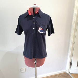 May include: A navy blue short-sleeved polo shirt with a red collar and a white embroidered penguin on the pocket.