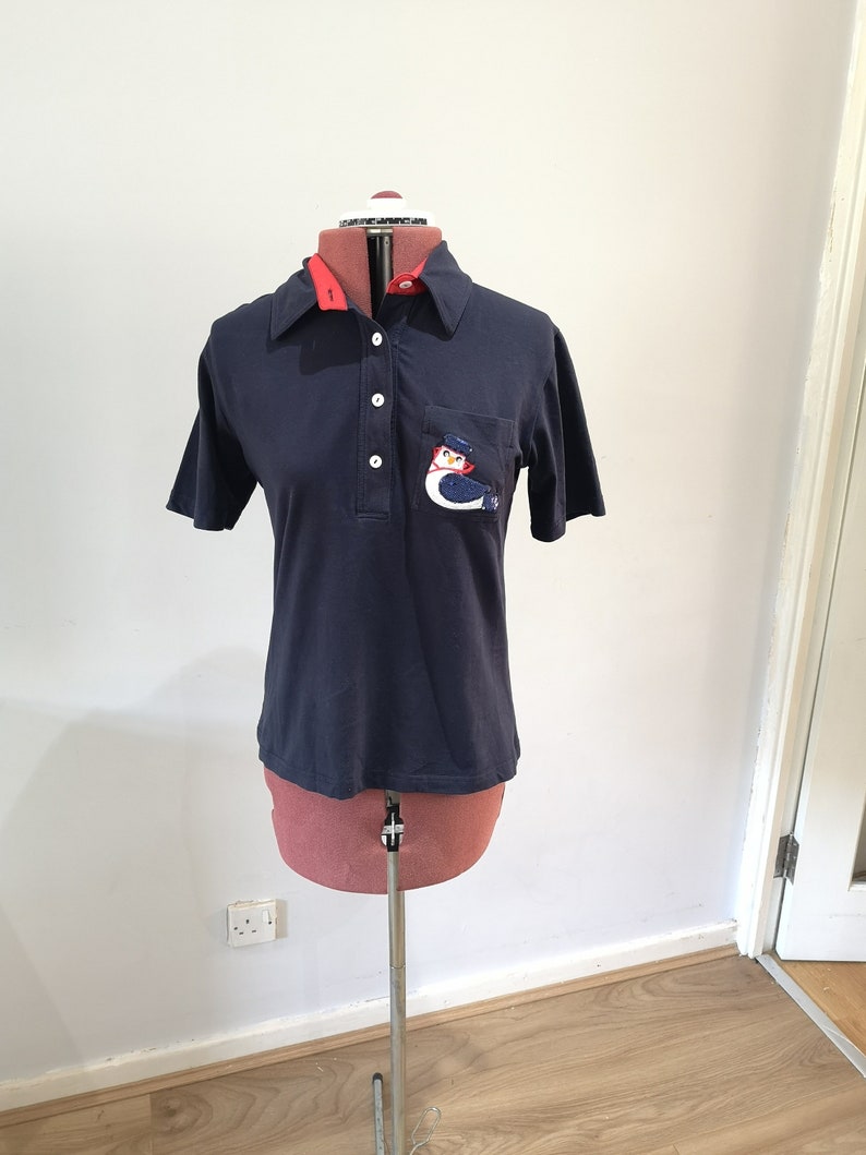 May include: A navy blue short-sleeved polo shirt with a red collar and a white embroidered penguin on the pocket.