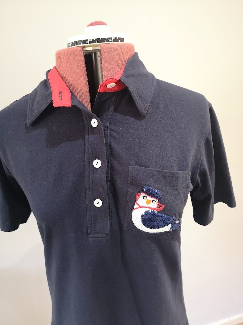 May include: A navy blue polo shirt with a red collar and a white button placket. The shirt has a pocket on the left side with a sequined penguin design.