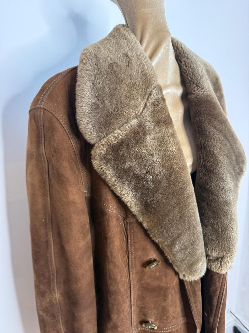 May include: A brown suede coat with a tan faux fur collar. The coat features a double-breasted design with two wooden toggle closures. The coat is displayed on a mannequin.