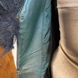 May include: Close-up of a dark green leather jacket with black fur trim. The jacket features gold-colored hook-and-eye closures. The interior lining is a lighter green, and the jacket appears to be well-constructed.