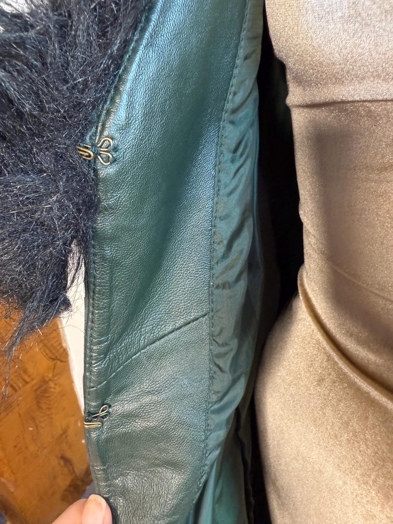 May include: Close-up of a dark green leather jacket with black fur trim. The jacket features gold-colored hook-and-eye closures. The interior lining is a lighter green, and the jacket appears to be well-constructed.