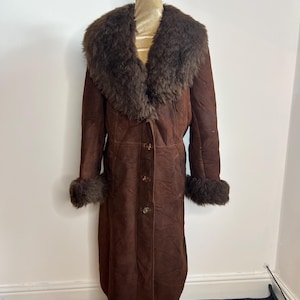 May include: A long, brown suede coat with a large, fluffy brown fur collar and cuffs. The coat has a button-up front and is displayed on a mannequin. A vintage, warm outerwear garment.