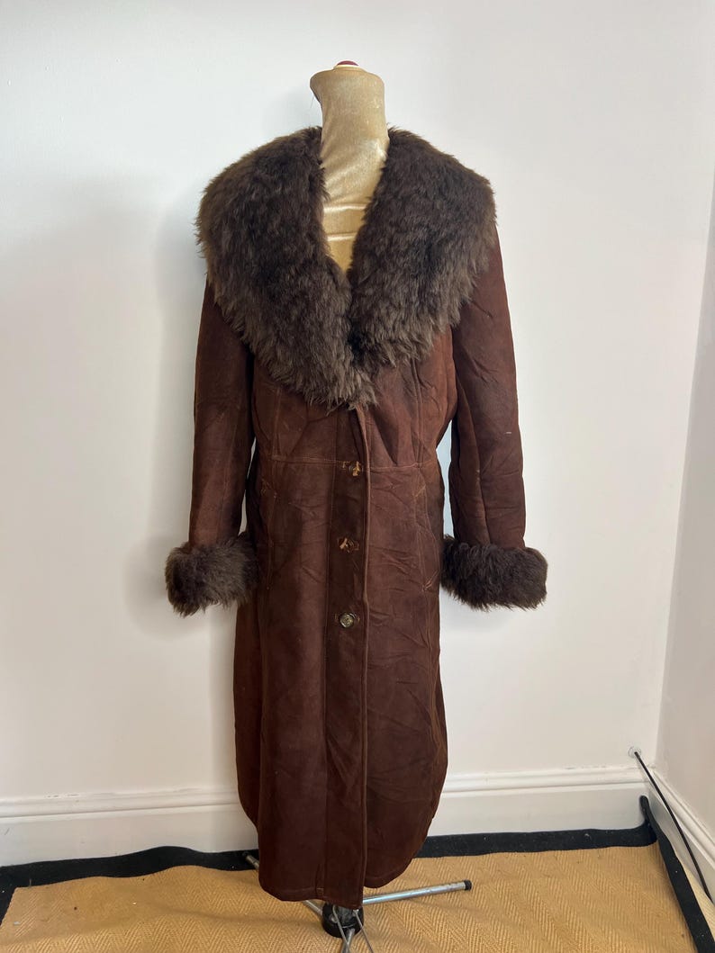 May include: A long, brown suede coat with a large, fluffy brown fur collar and cuffs. The coat has a button-up front and is displayed on a mannequin. A vintage, warm outerwear garment.