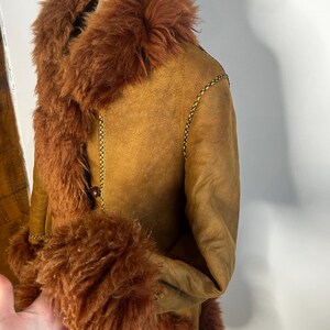 May include: A brown shearling coat with a faux fur collar and cuffs. The coat features a button closure and gold stitching details. The garment is displayed on a mannequin.
