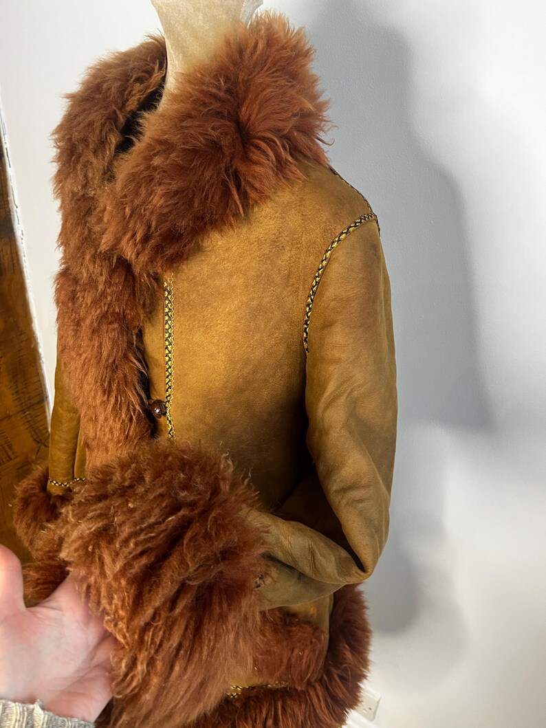 May include: A brown shearling coat with a faux fur collar and cuffs. The coat features a button closure and gold stitching details. The garment is displayed on a mannequin.