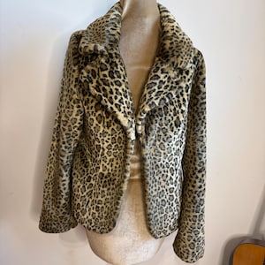 May include: A faux fur jacket featuring a brown, black, and tan leopard print pattern. The jacket has long sleeves, a wide notched collar, and a straight hemline. The garment is displayed on a mannequin, showcasing the textured, soft appearance of the material and the consistent animal print design across the front panels and collar.