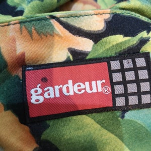 May include: A red and black rectangular label with the word "gardeur" in white lettering and a registered trademark symbol. The label is attached to a green and orange fabric with a floral pattern.