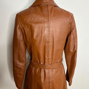 May include: A vintage brown leather coat with a belted waist and a classic collar. The coat has long sleeves and a tailored fit, showcasing a retro style. The leather appears to be well-maintained, with visible stitching details.