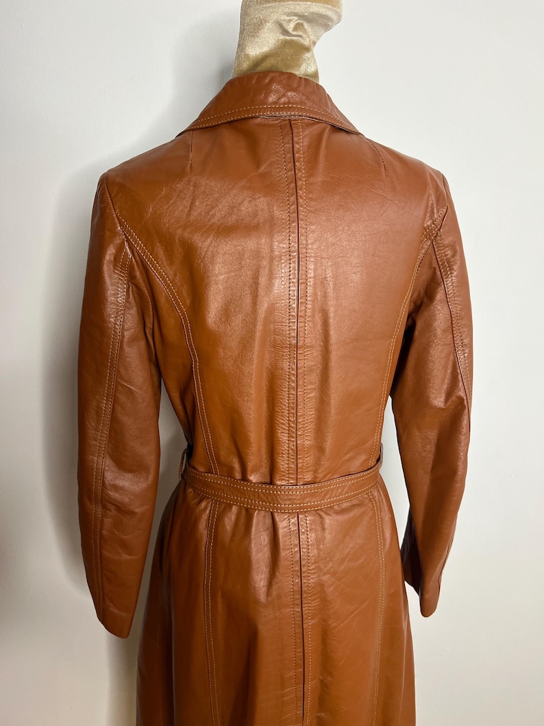 May include: A vintage brown leather coat with a belted waist and a classic collar. The coat has long sleeves and a tailored fit, showcasing a retro style. The leather appears to be well-maintained, with visible stitching details.