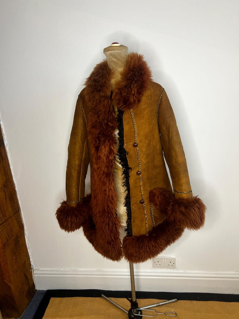 May include: A vintage brown suede coat with a faux fur collar, cuffs, and trim. The coat features a button-up front and decorative stitching. The coat is displayed on a mannequin.
