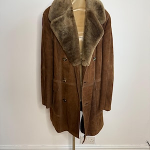 May include: A vintage brown suede coat with a shearling collar and lining. The coat features a double-breasted design with large buttons and a knee-length cut. The coat is displayed on a mannequin, highlighting its classic style and warm, textured appearance.