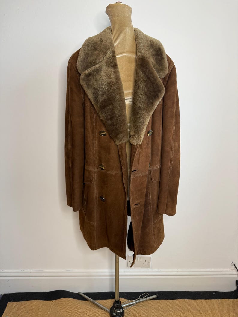 May include: A vintage brown suede coat with a shearling collar and lining. The coat features a double-breasted design with large buttons and a knee-length cut. The coat is displayed on a mannequin, highlighting its classic style and warm, textured appearance.
