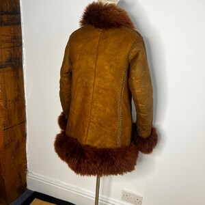 May include: A vintage brown shearling coat with a faux fur collar and cuffs. The coat is displayed on a mannequin, showcasing its warm, textured appearance. The coat features decorative stitching along the seams.