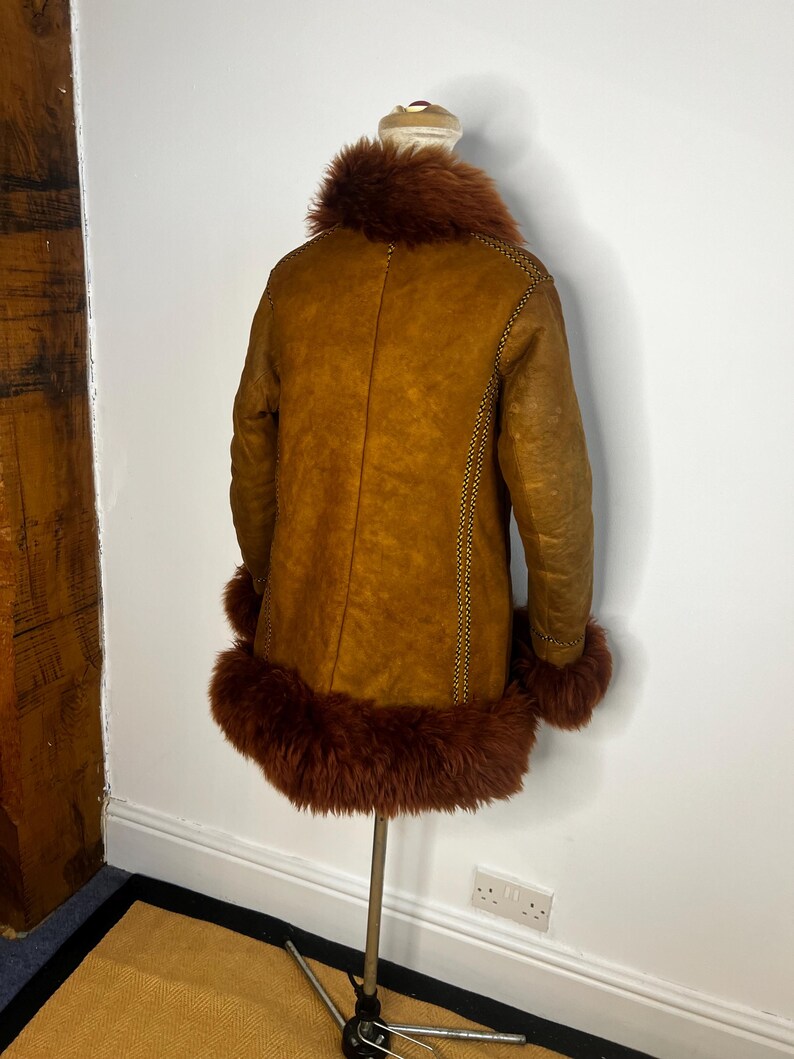 May include: A vintage brown shearling coat with a faux fur collar and cuffs. The coat is displayed on a mannequin, showcasing its warm, textured appearance. The coat features decorative stitching along the seams.