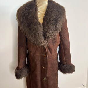 May include: A long, brown suede coat with a large, fluffy, dark brown fur collar and cuffs. The coat has a button-up front and is displayed on a mannequin. A vintage-style garment.