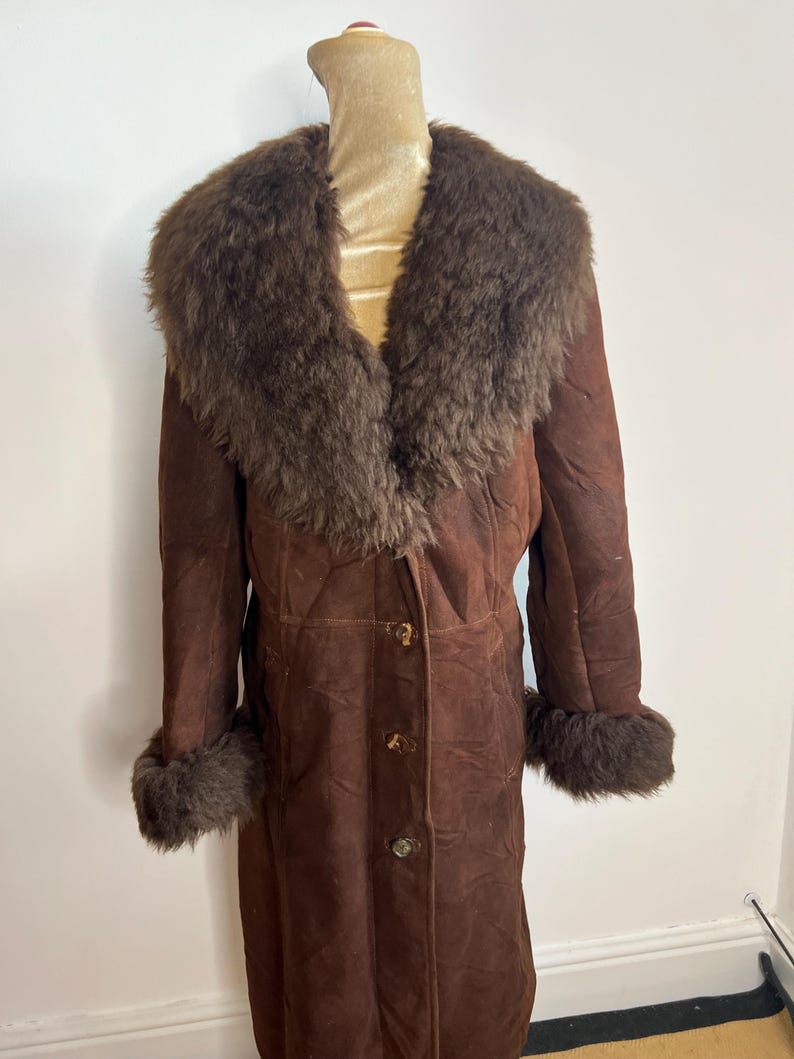 May include: A long, brown suede coat with a large, fluffy, dark brown fur collar and cuffs. The coat has a button-up front and is displayed on a mannequin. A vintage-style garment.