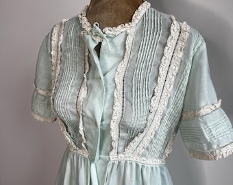 May include: A light blue vintage dress with short sleeves. The dress features vertical pleats on the bodice, accented with white lace trim. The neckline and sleeve cuffs are also trimmed with lace. The dress is displayed on a mannequin.