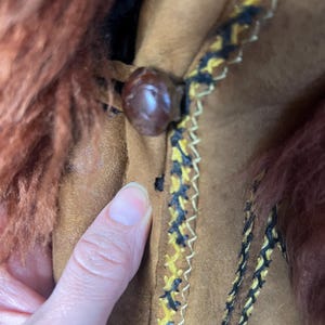 May include: Close-up of a brown suede jacket with a fur collar. The jacket features a decorative trim with yellow and black stitching. A large, round, brown button fastens the jacket.