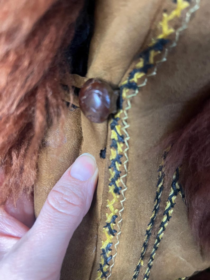 May include: Close-up of a brown suede jacket with a fur collar. The jacket features a decorative trim with yellow and black stitching. A large, round, brown button fastens the jacket.