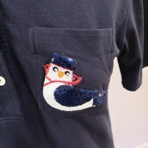 May include: A navy blue polo shirt with a patch pocket featuring a white and blue penguin with red accents. The penguin is made of sequins.