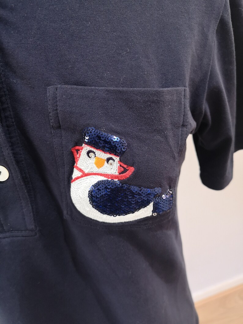 May include: A navy blue polo shirt with a patch pocket featuring a white and blue penguin with red accents. The penguin is made of sequins.