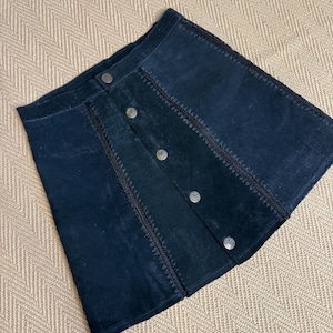 以下が含まれることがあります： ボタン留めと装飾的なステッチが施された、濃い青色のスエードスカート。 スカートはクラシックなAラインの形で、シルバー色のボタンが付いています。 ステッチのディテールが、衣服に質感と視覚的な面白さを加えています。