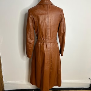 May include: A vintage brown leather trench coat with a belted waist and long sleeves. The coat is displayed on a mannequin, showcasing its tailored fit and classic design. The coat's color is a warm, rich brown.